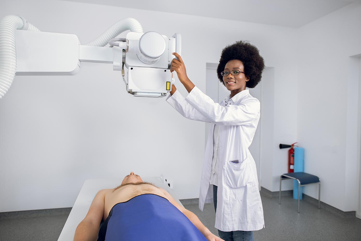 Smiling professional doctor in uniform posing to camera, while taking chest Xray of male Caucasian patient. X-ray scan of lungs and skeleton in modern clinic